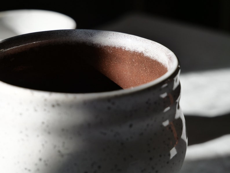 May include: Close-up of a speckled white ceramic mug with a textured exterior. The interior is a warm brown color. The rim has a white and dark speckled pattern. The mug is partially in shadow.