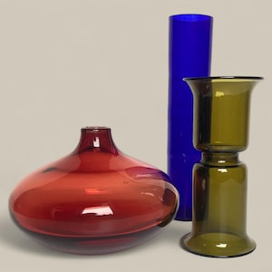 May include: Three glass vases in shades of red, blue, and green. The red vase is rounded, the blue is a tall cylinder, and the green is a two-part design. The vases are decorative.