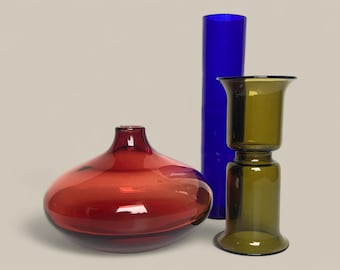 May include: Three glass vases in shades of red, blue, and green. The red vase is rounded, the blue is a tall cylinder, and the green is a two-part design. The vases are decorative.