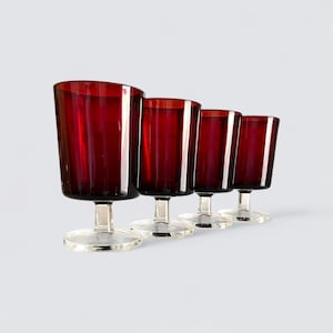 Könnte beinhalten: Vier Weingläser mit Stiel in tiefroter Farbe. Die Gläser haben einen klaren, kreisförmigen Fuß und einen kurzen Stiel. Die Glasschalen sind rechteckig mit vertikalen Linien. Die Gläser sind in einer Reihe angeordnet, leicht abgewinkelt.