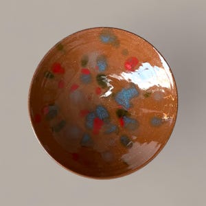 Op de afbeelding: Een handgemaakte keramische kom met een gespikkeld ontwerp. De kom is warmbruin van kleur met rode, blauwe en groene spikkels. De binnenkant van de kom is glanzend en reflecteert licht. De kom is rond en heeft een eenvoudige, elegante vorm.