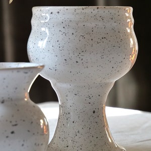 May include: A speckled white ceramic vase with a wide, rounded top and a pedestal base. The vase has a textured surface with small, dark gray speckles. The design is reminiscent of a chalice or goblet. A smaller, similar vase is partially visible in the foreground.