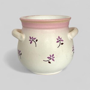 Peut inclure: Un pot en céramique blanche avec un bord rose et des motifs de fleurs violettes. Le pot a deux anses.