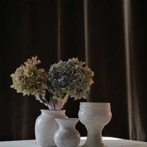 May include: Three speckled white ceramic vases of varying sizes are arranged on a white surface. One vase holds a bouquet of dried hydrangeas in shades of green, blue, and pink. The vases have a speckled pattern.