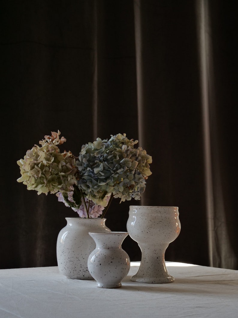 May include: Three speckled white ceramic vases of varying sizes are arranged on a white surface. One vase holds a bouquet of dried hydrangeas in shades of green, blue, and pink. The vases have a speckled pattern.
