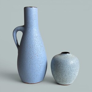 Könnte beinhalten: Eine hohe, hellblaue Keramikvase mit seitlichem Henkel und texturierter, gesprenkelter Oberfläche, neben einer kleineren, runden, hellblau-weiß gesprenkelten Keramikvase. Beide Gefäße haben einen dunkelbraunen Rand und eine raue, körnige Struktur.