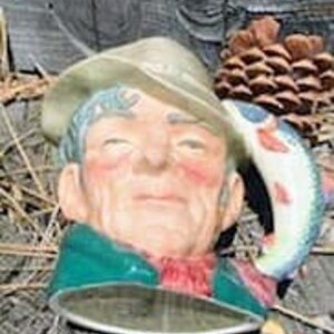 May include: Ceramic mug shaped like a fisherman's head, wearing a hat and green jacket. The handle is a fish with red and white markings. The mug is set against a rustic wooden background with pinecones.