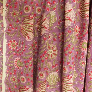 May include: A close-up of a fabric with a floral pattern. The fabric has a lavender background with a repeating design of flowers and leaves in shades of red, green, and pink. The edges of the fabric are visible.