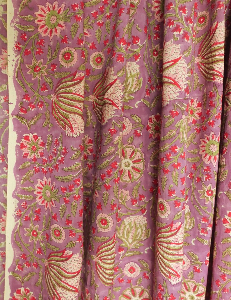 May include: A close-up of a fabric with a floral pattern. The fabric has a lavender background with a repeating design of flowers and leaves in shades of red, green, and pink. The edges of the fabric are visible.