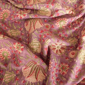 May include: A close-up of a fabric with a floral pattern. The fabric is a muted purple color with a repeating design of flowers in red, green, and cream. The fabric is draped, showing folds and texture.