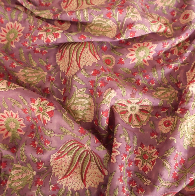 May include: A close-up of a fabric with a floral pattern. The fabric is a muted purple color with a repeating design of flowers in red, green, and cream. The fabric is draped, showing folds and texture.