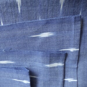 May include: Close-up of blue fabric with white, horizontal, brushstroke-like patterns. The fabric is folded, revealing a pocket. The texture of the fabric is visible, with a woven appearance. The overall aesthetic is simple and elegant.