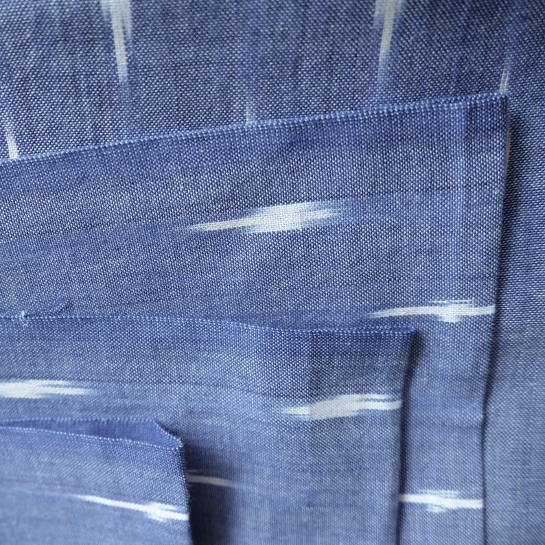 May include: Close-up of blue fabric with white, horizontal, brushstroke-like patterns. The fabric is folded, revealing a pocket. The texture of the fabric is visible, with a woven appearance. The overall aesthetic is simple and elegant.