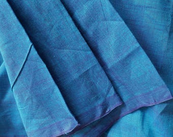 May include: A piece of teal blue fabric with a visible woven texture and subtle horizontal lines. The material is folded, creating layered sections that show the depth of the blue colour and the natural weave of the textile fibres throughout.