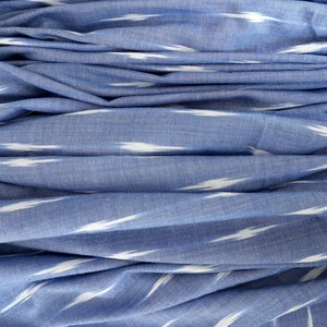 May include: Close-up of blue fabric with white ikat-style stripes. The fabric is folded, creating a layered effect. The material appears soft and lightweight, suitable for clothing or home decor.