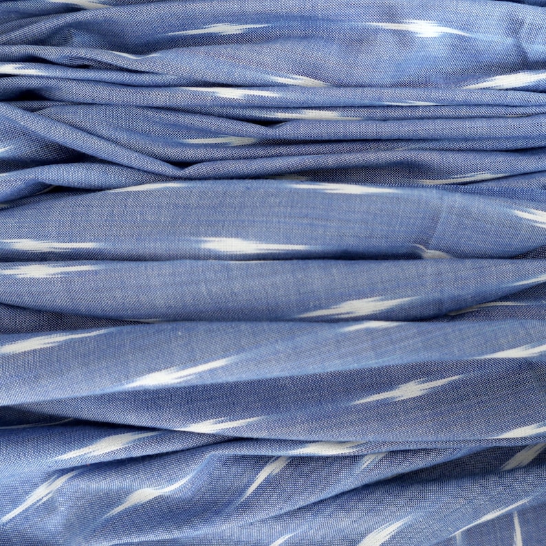 May include: Close-up of blue fabric with white ikat-style stripes. The fabric is folded, creating a layered effect. The material appears soft and lightweight, suitable for clothing or home decor.