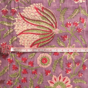 May include: Close-up of a floral fabric with a measuring tape. The fabric features a large, stylized flower in shades of pink, red, and green on a lavender background. The measuring tape shows measurements in inches.