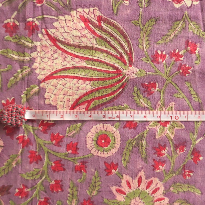 May include: Close-up of a floral fabric with a measuring tape. The fabric features a large, stylized flower in shades of pink, red, and green on a lavender background. The measuring tape shows measurements in inches.