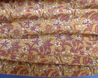 May include: Close-up of a fabric with a floral pattern. The fabric has a mustard yellow background with white flowers and maroon accents. The flowers are daisy-like with detailed petals and surrounded by maroon leaves.