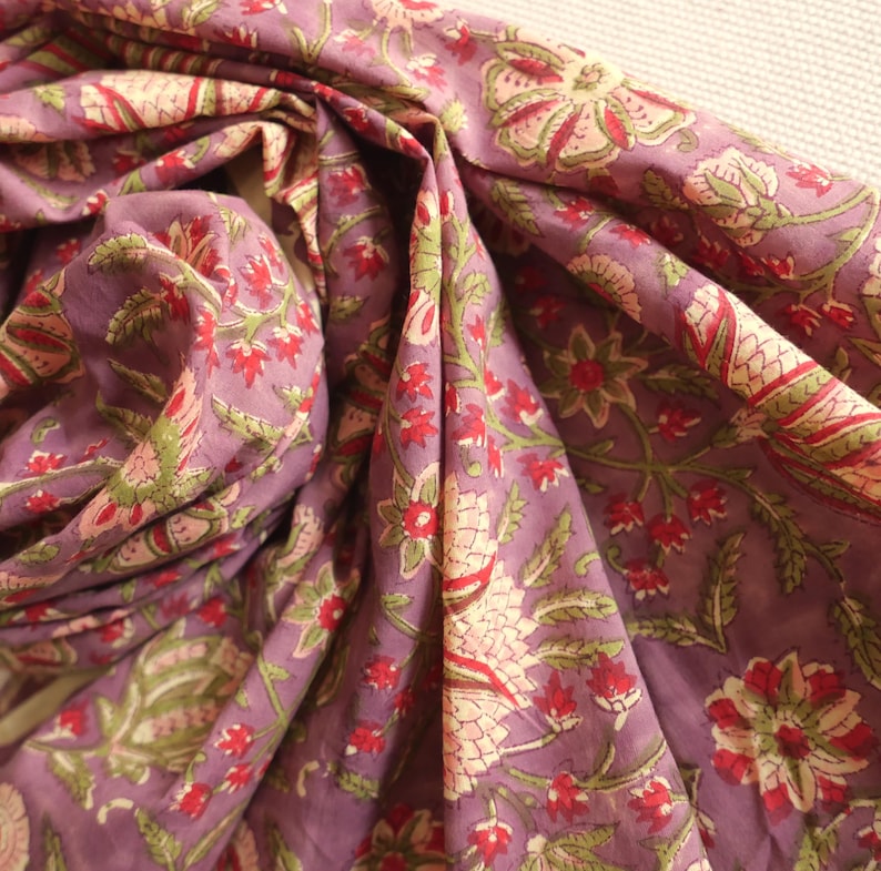 May include: A draped fabric with a floral pattern in shades of purple, red, green, and cream. The fabric is soft and flowing, with visible folds and creases. The floral design features stylized flowers and leaves.