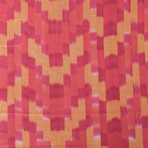 Op de afbeelding: Close-up van een stof met een geometrisch patroon. Het ontwerp bevat verticale en horizontale blokken in tinten rood, roze en oranje. De stof heeft een getextureerd uiterlijk, wat wijst op een geweven materiaal. De algehele esthetiek is levendig en kleurrijk.