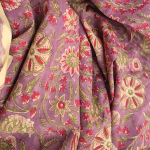 May include: Close-up of a purple fabric with a floral pattern. The design features large flowers in pink and green, with red accents, and green leaves. The fabric is draped, showing the texture and folds.