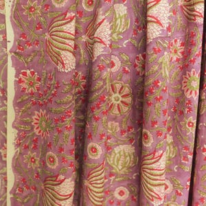 May include: Close-up of a fabric with a floral pattern. The fabric features a repeating design of flowers and leaves in shades of red, green, and cream on a lavender background. The fabric is likely used for curtains or upholstery.