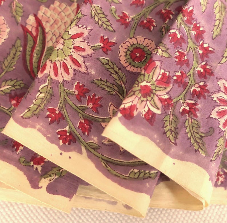 May include: Close-up of a folded fabric with a floral pattern. The fabric is a muted purple color with a cream-colored border. The floral design features red and pink flowers with green leaves.