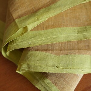 Könnte beinhalten: Nahaufnahme von gefaltetem Stoff mit beigefarbener Basis und hellgrünen Rändern. Der Stoff hat ein strukturiertes Aussehen mit sichtbaren Fäden und einer leicht zerknitterten Textur. Die grünen Ränder setzen Farbakzente.