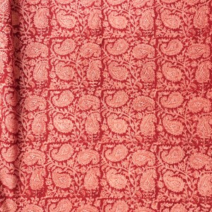 Könnte beinhalten: Ein Textilstoff mit einem sich wiederholenden Muster aus hellrosa Paisley-Motiven und komplizierten Blumenranken vor einem tiefroten Hintergrund. Das Design ist in einem Raster aus quadratischen Abschnitten angeordnet und zeigt einen botanischen Druck.