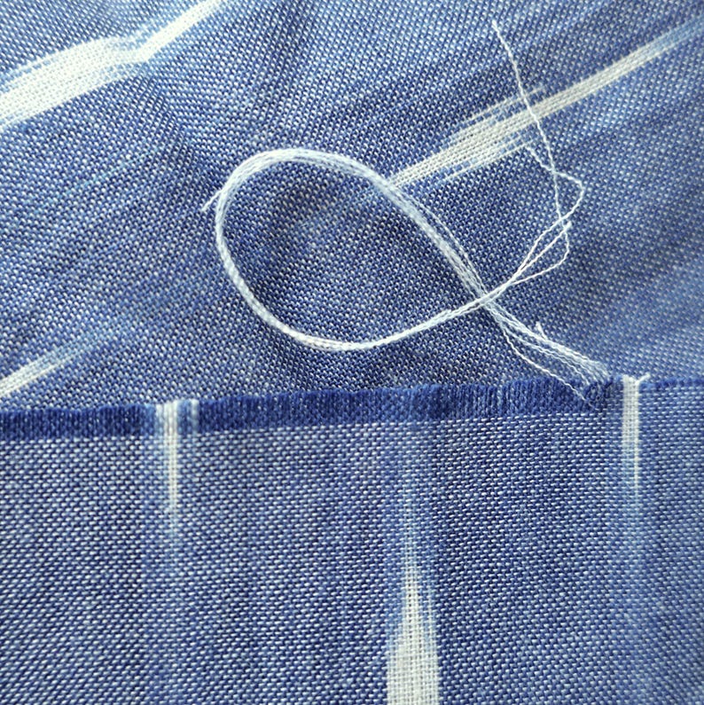 May include: Close-up of blue fabric with white abstract streaks. A loop of white thread is visible, along with a frayed edge. The fabric has a textured appearance, with a darker blue border along the bottom.