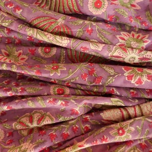 May include: Close-up of a folded fabric with a floral pattern. The fabric is a muted purple color with a repeating design of red, green, and off-white flowers and leaves. The fabric is soft and appears to be cotton or linen.