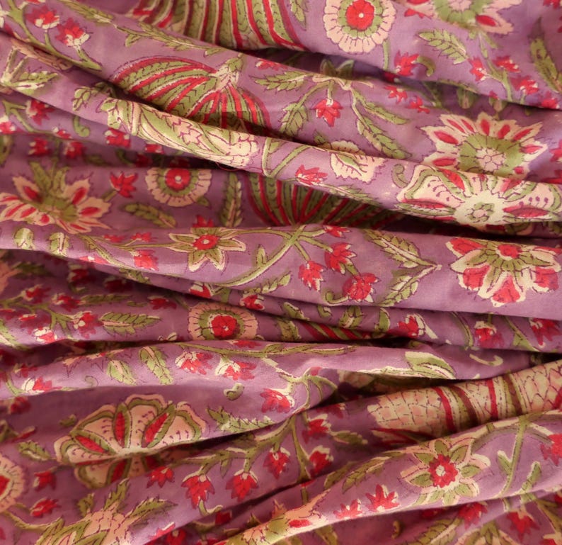 May include: Close-up of a folded fabric with a floral pattern. The fabric is a muted purple color with a repeating design of red, green, and off-white flowers and leaves. The fabric is soft and appears to be cotton or linen.