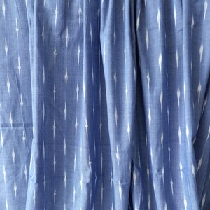 May include: Close-up of blue fabric with a white ikat pattern. The fabric is draped, showing folds and texture. The pattern consists of vertical white dashes against a solid blue background, creating a textured effect.