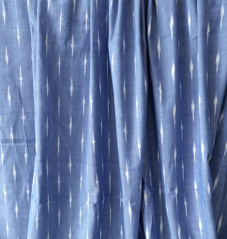 May include: Close-up of blue fabric with a white ikat pattern. The fabric is draped, showing folds and texture. The pattern consists of vertical white dashes against a solid blue background, creating a textured effect.