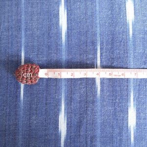 May include: A small, heart-shaped, crocheted item in shades of red and grey, next to a measuring tape. The tape shows measurements in centimetres, with the item at the zero mark. The background is a blue fabric with white vertical stripes.