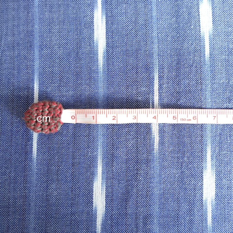 May include: A small, heart-shaped, crocheted item in shades of red and grey, next to a measuring tape. The tape shows measurements in centimetres, with the item at the zero mark. The background is a blue fabric with white vertical stripes.