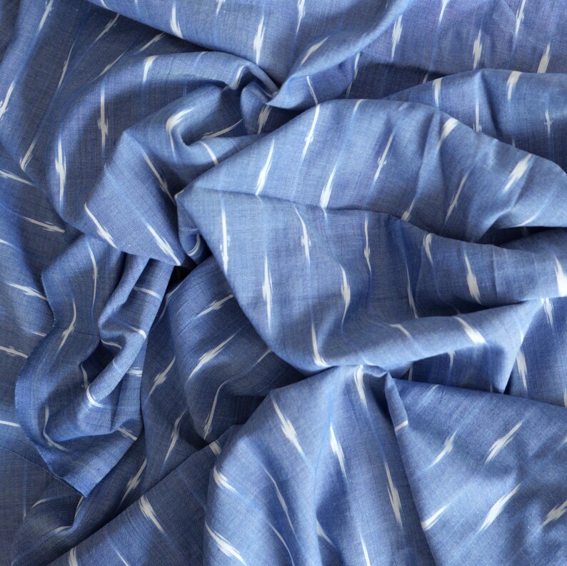 May include: A close-up shot of blue fabric with a white ikat pattern. The fabric is creased, creating shadows and highlights. The pattern features white, vertical, brushstroke-like designs against a blue backdrop. The fabric appears to be cotton or a similar material.