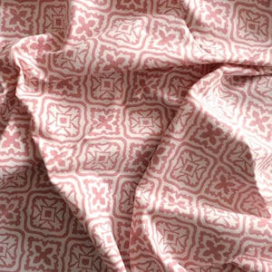 May include: A close-up of a fabric with a repeating geometric pattern in light pink and white. The design features square and floral shapes, creating a textured appearance. The fabric is draped, showing folds and shadows.