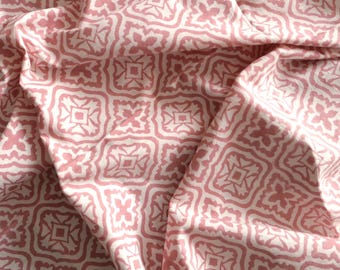 May include: A close-up of a fabric with a repeating geometric pattern in light pink and white. The design features square and floral shapes, creating a textured appearance. The fabric is draped, showing folds and shadows.