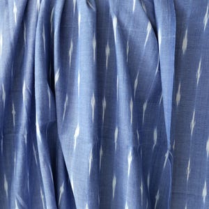 May include: Close-up of blue fabric with a white ikat pattern. The fabric is draped, showing folds and texture. The pattern consists of vertical white dashes against a blue background, creating a textured effect. The fabric appears to be cotton or a similar material.