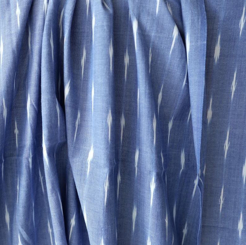May include: Close-up of blue fabric with a white ikat pattern. The fabric is draped, showing folds and texture. The pattern consists of vertical white dashes against a blue background, creating a textured effect. The fabric appears to be cotton or a similar material.