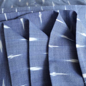 May include: Close-up of blue fabric with white, horizontal, brushstroke-like patterns. The fabric is folded, showing the texture and weave. The colour is a medium blue, and the white patterns create a contrasting design. This textile is suitable for various sewing projects.