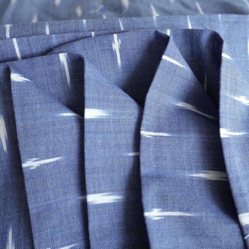 May include: Close-up of blue fabric with white, horizontal, brushstroke-like patterns. The fabric is folded, showing the texture and weave. The colour is a medium blue, and the white patterns create a contrasting design. This textile is suitable for various sewing projects.