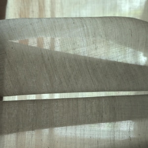 Könnte beinhalten: Nahaufnahme eines hellbeigen Leinenstoffs, möglicherweise ein Vorhang oder eine Gardine, mit sichtbarer Textur und subtilen Schatten. Der Stoff ist gefaltet, wodurch Tiefe entsteht und das Gewebe hervorgehoben wird. Die Gesamtästhetik ist weich und natürlich.