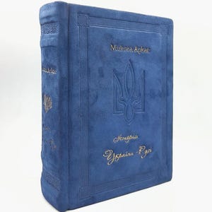 May include: A blue book with gold lettering and a Ukrainian coat of arms. The book's cover features the title "Микола Аркас" and the words "Історія України-Руси". The book has a textured, suede-like appearance.
