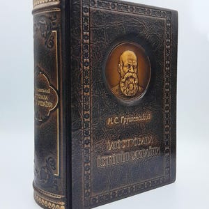 Puede incluir: Un libro encuadernado en cuero marrón oscuro con acabado texturizado y letras doradas. La portada presenta un retrato circular en relieve de un hombre barbudo, con el texto M.C. Grushevsky e Historia ilustrada de Ucrania en ucraniano.