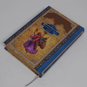 May include: Hardcover book with a blue spine and cover accents. The cover features a colorful illustration of a woman riding a creature, with text in a blue banner. A cream-colored ribbon bookmark is visible.