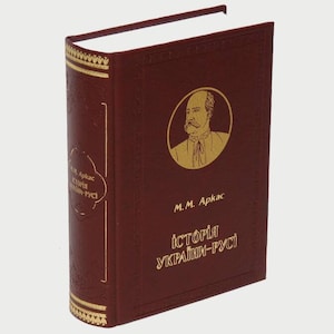 May include: A burgundy leather-bound book with gold accents and a portrait of a man with a beard and mustache on the cover. The title of the book is "Історія України-Руси" by M.M. Arkac.