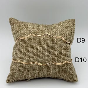 以下が含まれることがあります： 波状の縁を持つ2つの金色のブレスレットが、ジュートの枕の上に展示されています。ブレスレットは細く繊細で、シンプルでエレガントなデザインです。ブレスレットにはD9とD10のラベルが付いています。